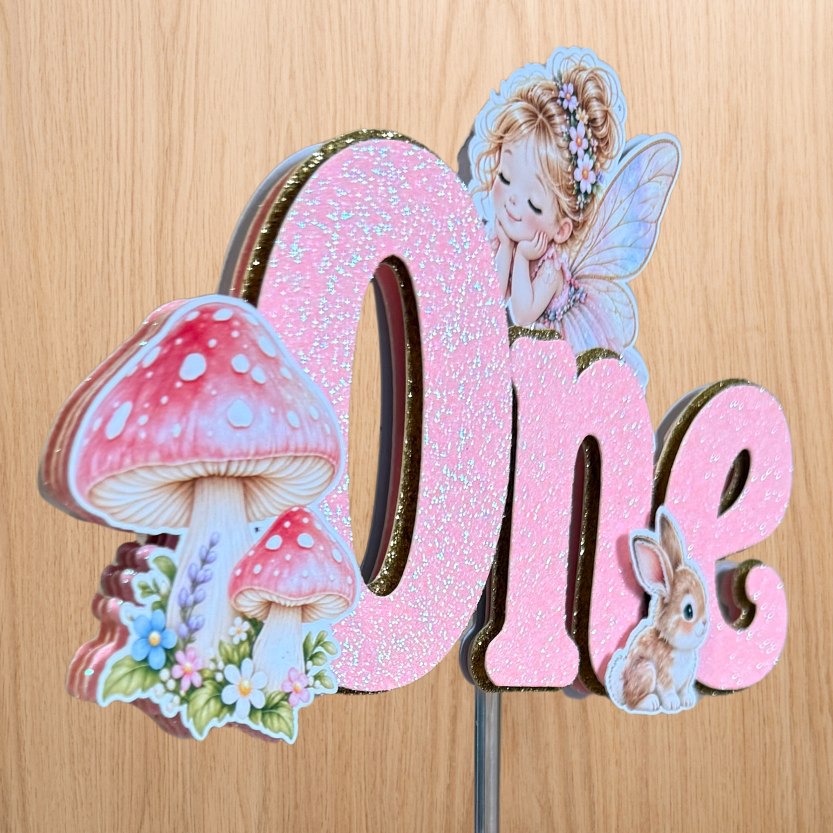 Handmade layered fairy garden first birthday cake topper showing dimensional glitter lettering